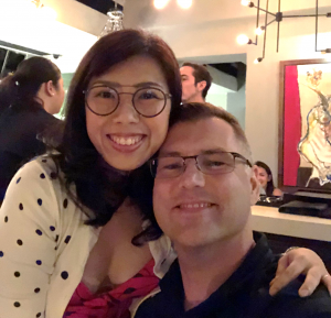 Woman with dark hair and glasses poses with a man with short light hair and glasses. Both are smiling.