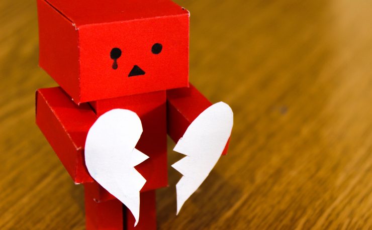 Photo of a red toy robot with a tear drawn onto its face holding two halves of a broken heart in its hands.