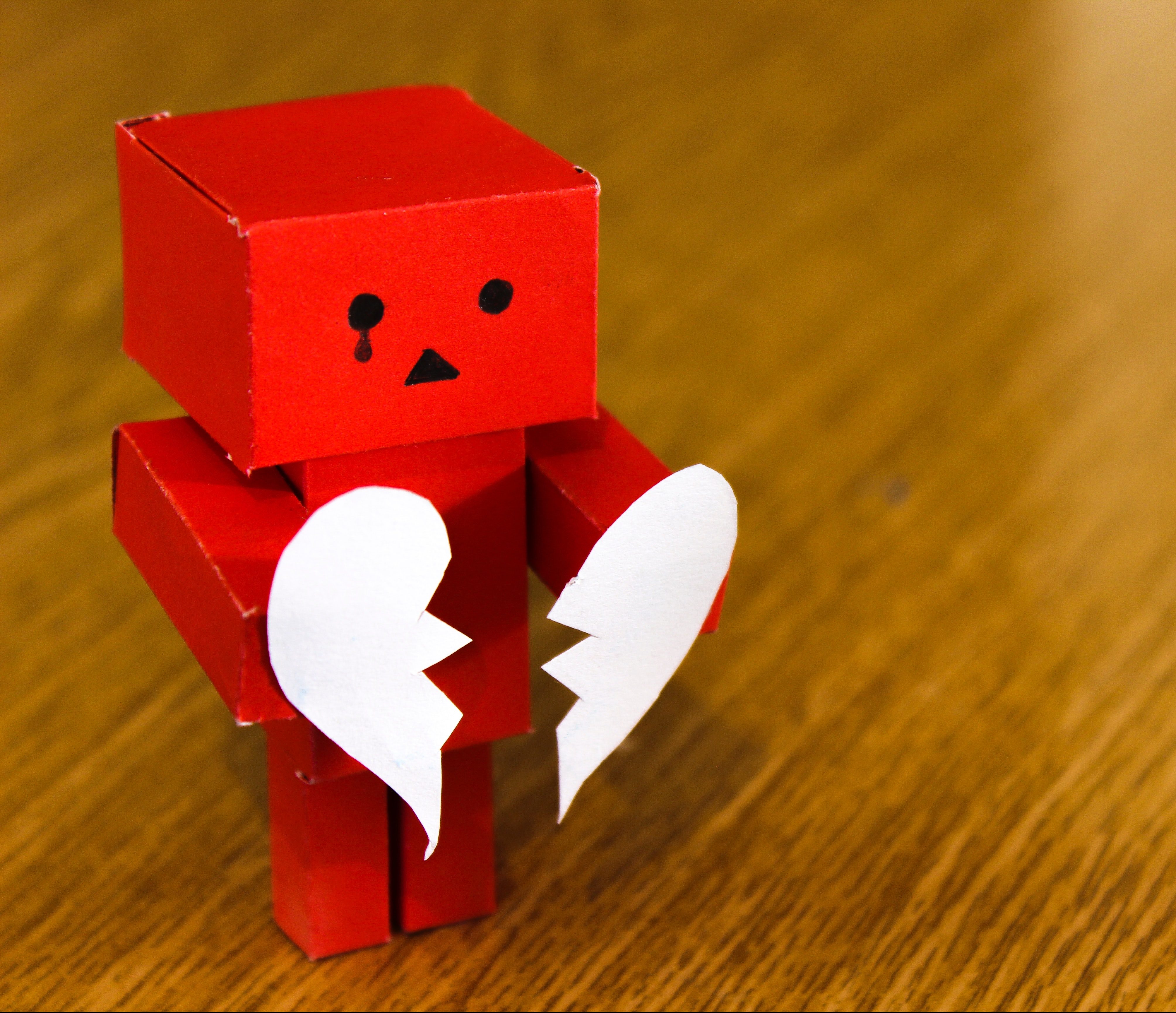 Photo of a red toy robot with a tear drawn onto its face holding two halves of a broken heart in its hands.