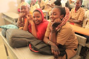 Three female students sitting in class with their backpacks and smiling.